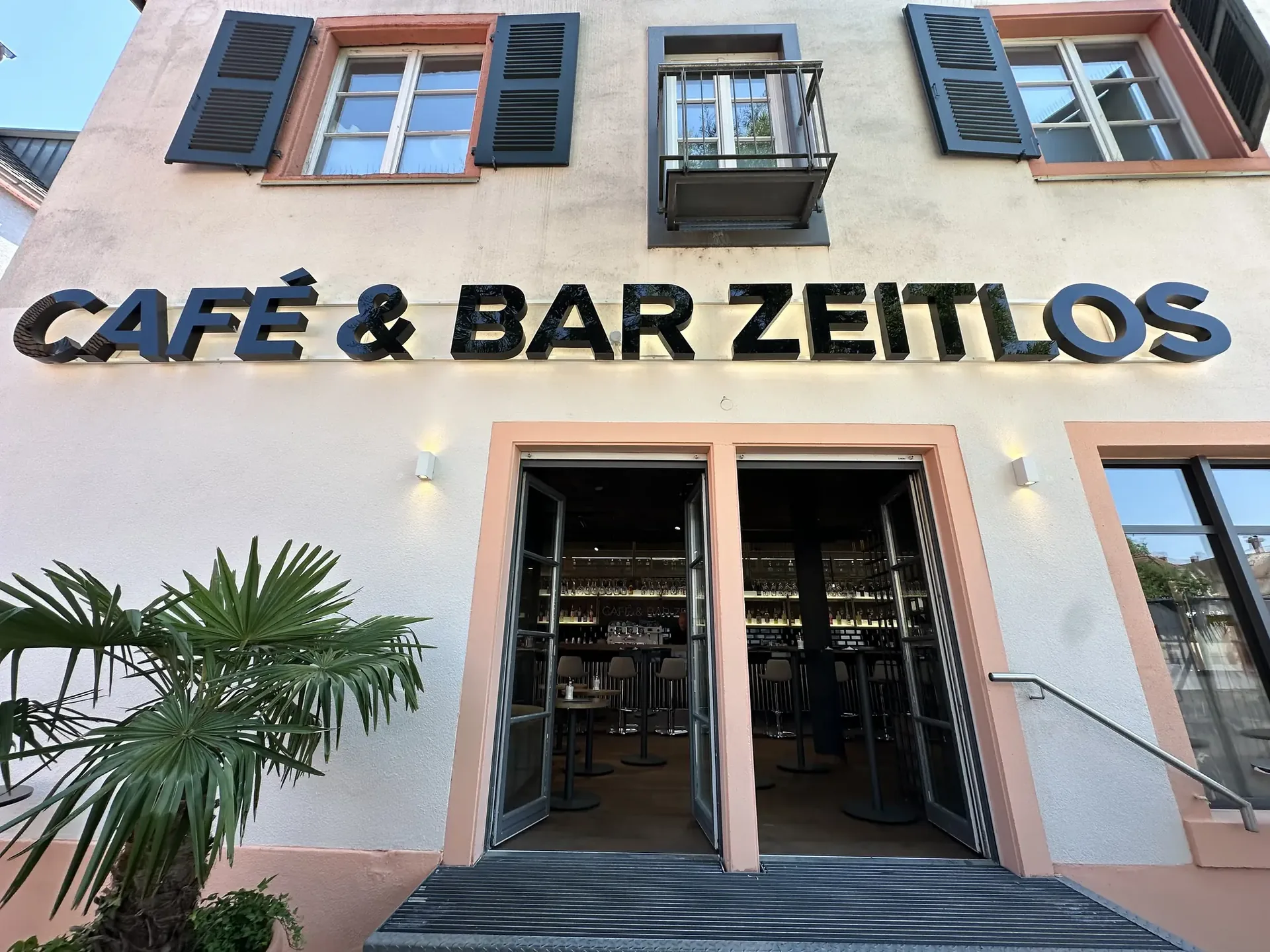 Fassade der Café & Bar Zeitlos mit Leuchtschrift, Eingang und Palme