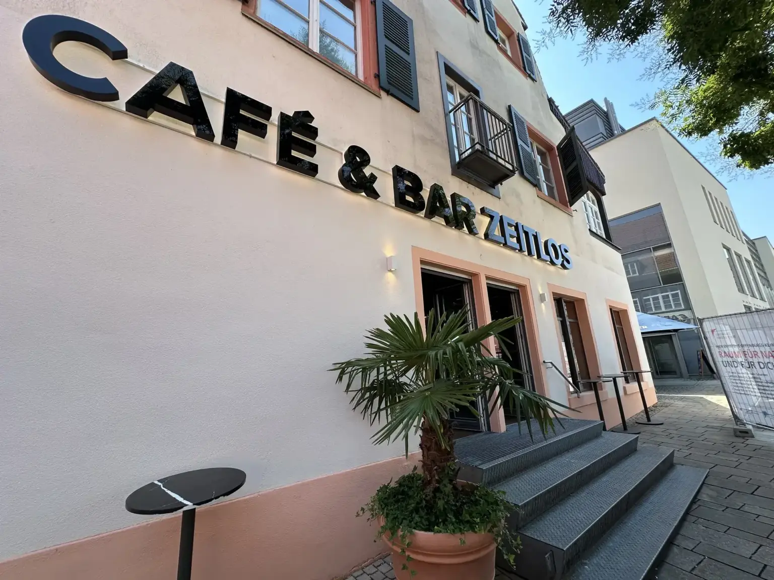 Hausfassade mit Schriftzug „Café & Bar Zeitlos“ und Stehtisch im Freien